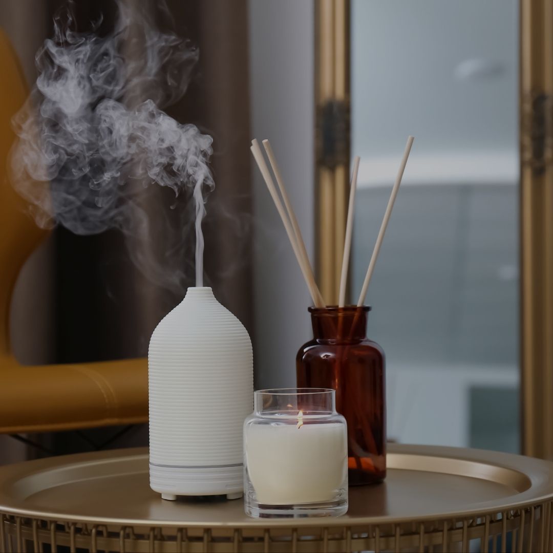 A white ribbed diffuser with mist coming out, styled on a round gold tray next to a white candle in a glass jar and a reed diffuser in an amber glass bottle.