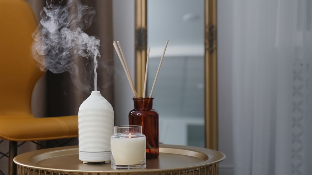 A white diffuser with misting steam sits on a round gold tray next to a brown reed diffuser bottle and a small white candle in a glass jar, set in a modern interior with a yellow chair and mirror in the background.