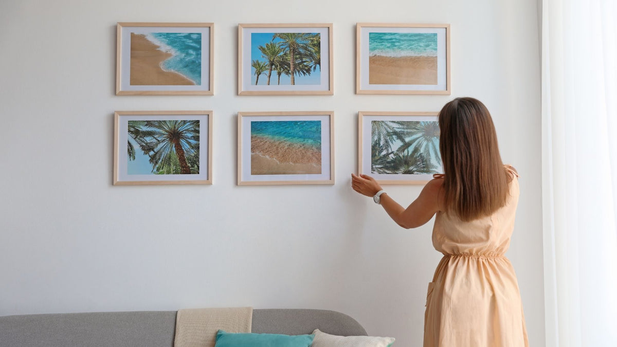 Woman arranging six framed coastal prints above a living room sofa